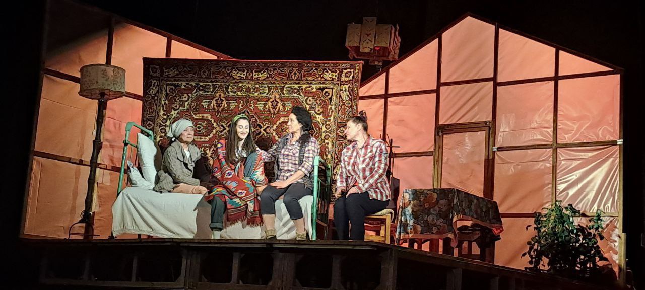 Сцена з театральної вистави, де чотири жінки сидять і спілкуються. На задньому плані — стилізовані декорації, що нагадують стіни хат, на одній з яких висить великий східний килим.  На сцені — ліжко, стільці, абажур та побутові предмети. Жінки одягнені у народному або простому сільському стилі. Сцена яскраво освітлена, що підкреслює театральну атмосферу.