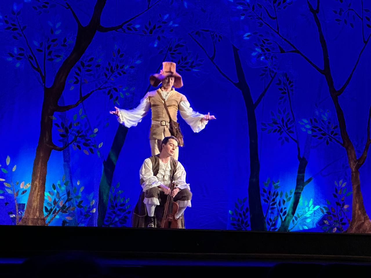 Сцена з театральної або оперної вистави: двоє чоловіків у сценічних костюмах, один з яких стоїть на плечах іншого. Фон — яскраво-синій із силуетами дерев, що створює атмосферу казкового лісу.  Чоловік знизу присідає і тримає віолончель, одягнений у білу сорочку та коричневий камзол. Чоловік зверху стоїть із розведеними руками, у високому капелюсі та коричневому одязі. Яскраве синє освітлення підкреслює декорації.