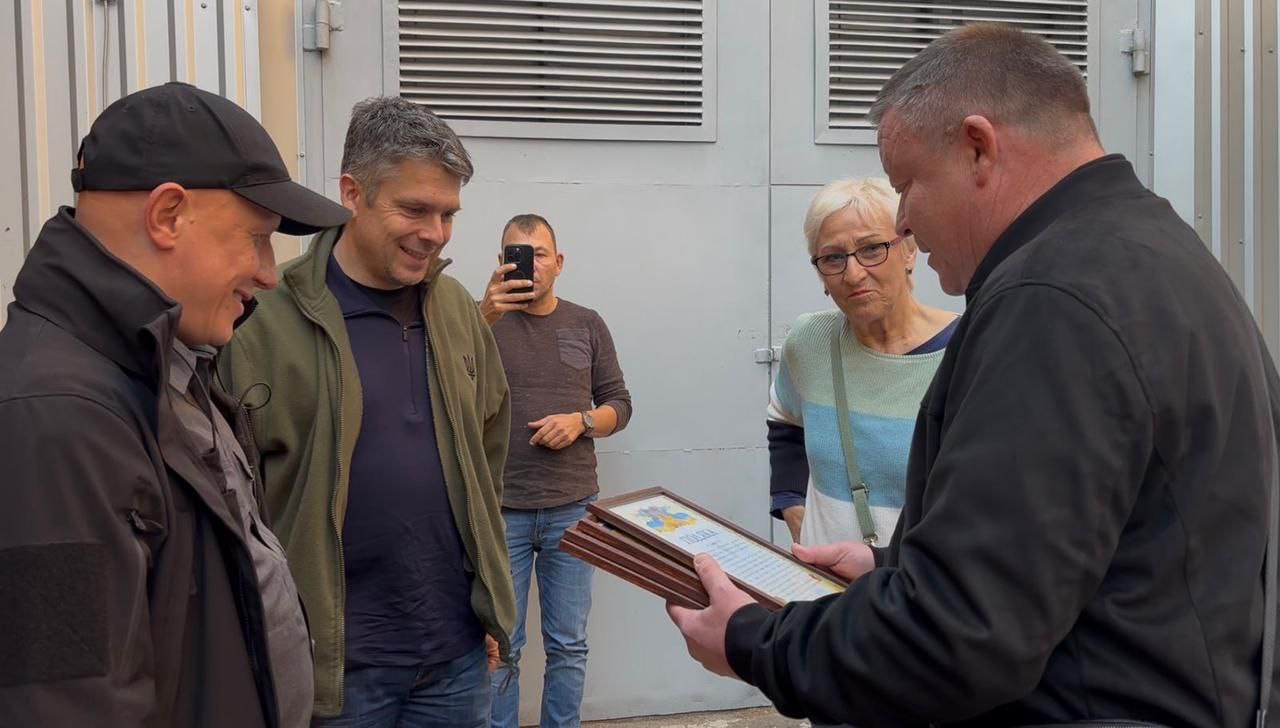 Група з чотирьох людей, включаючи жінку та трьох чоловіків, стоїть на вулиці. Чоловік на передньому плані, повернутий спиною, тримає стопку рамок з Подяками. Двоє чоловіків ліворуч посміхаються. На задньому плані інший чоловік фотографує подію на телефон.