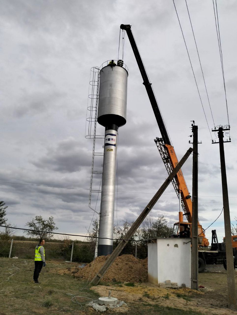 Монтаж металевої водонапірної вежі, де верхній бак підіймає стріловий кран. Біля основи вежі стоїть робітник у світловідбивному жилеті. Хмарний день. На фото видно робітників, кран та нову водонапірну вежу з написом UGM на циліндричній основі, що свідчить про будівельні або ремонтні роботи.