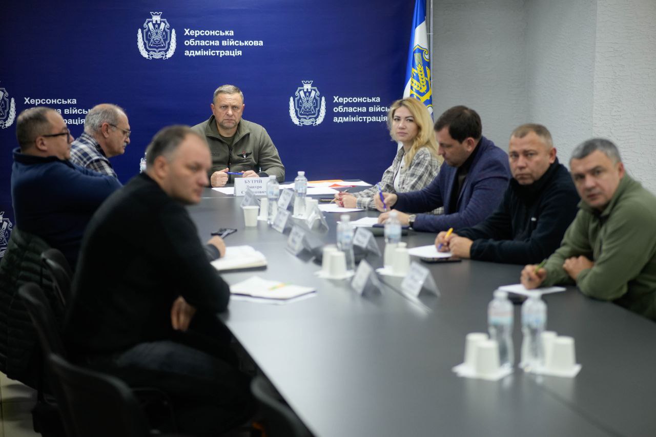Група з семи осіб, включаючи чоловіків і одну жінку, сидять за довгим столом на нараді в приміщенні Херсонської обласної військової адміністрації. Центральний учасник, одягнений у військову кофту - очолює зустріч. Фон — синя стіна з логотипами адміністрації та прапором України.
