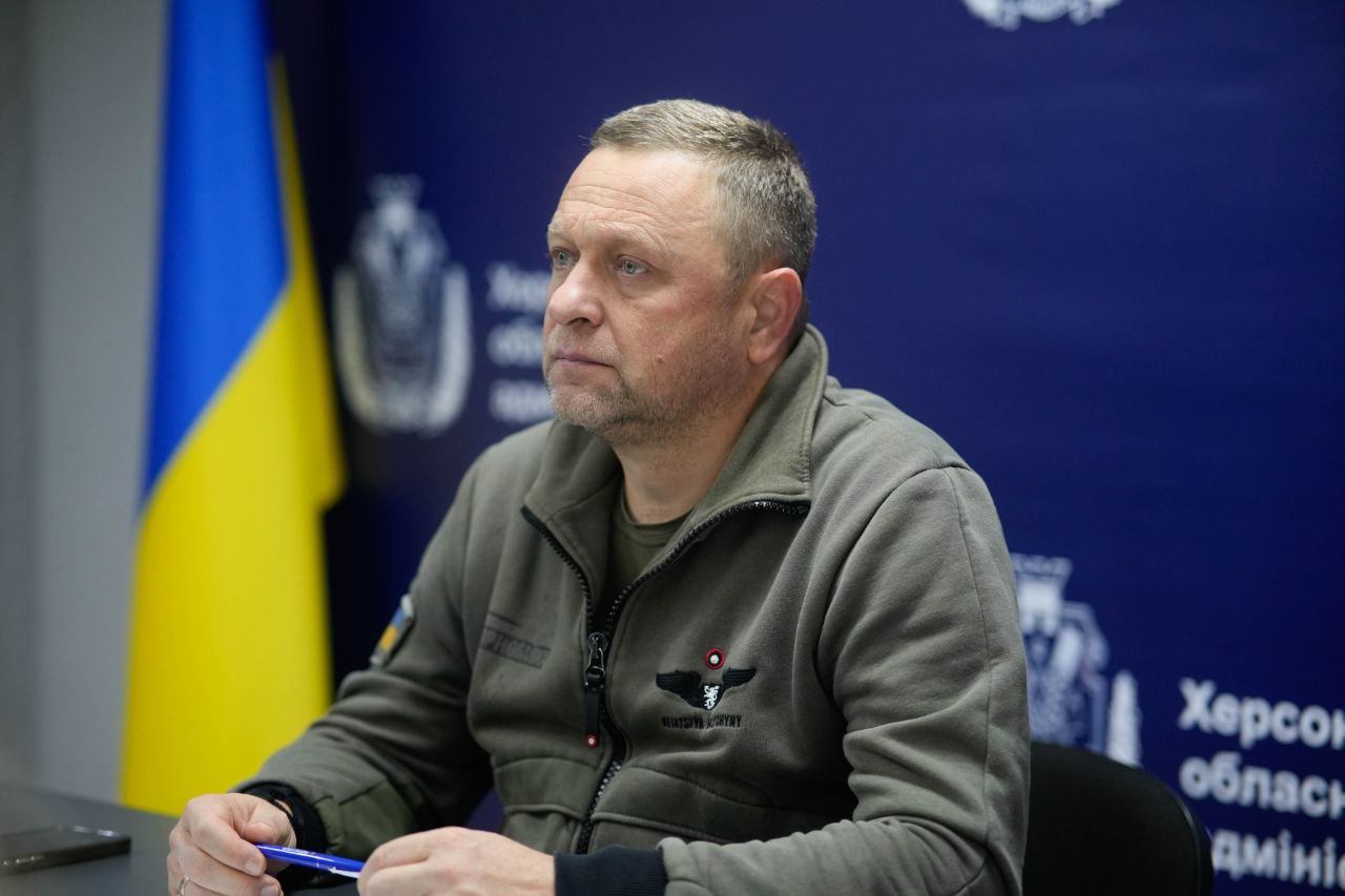 Чоловік середніх років у військовій флісовій кофті сидить за столом, уважно дивлячись убік. За його спиною видно прапор України та логотип Херсонської обласної військової адміністрації.