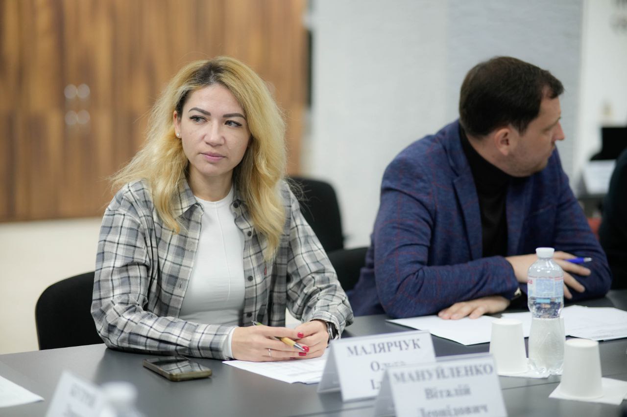 Жінка зі світлим волоссям сидить за столом під час наради, дивлячись убік. Вона одягнена у світлий лонгслів та картату сорочку. Перед нею стоять таблички з іменами учасників. Праворуч від неї сидить чоловік у темно-синьому піджаку.