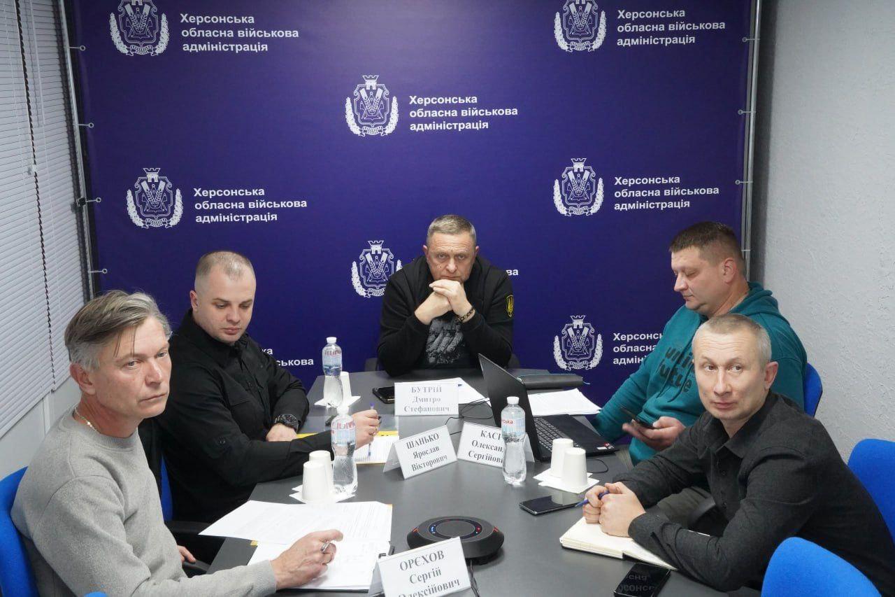 П'ятеро чоловіків сидять за столом під час наради у приміщенні Херсонської обласної військової адміністрації. На стіні за ними — емблема адміністрації. На столі видно пляшки з водою, папери та імена учасників зустрічі.