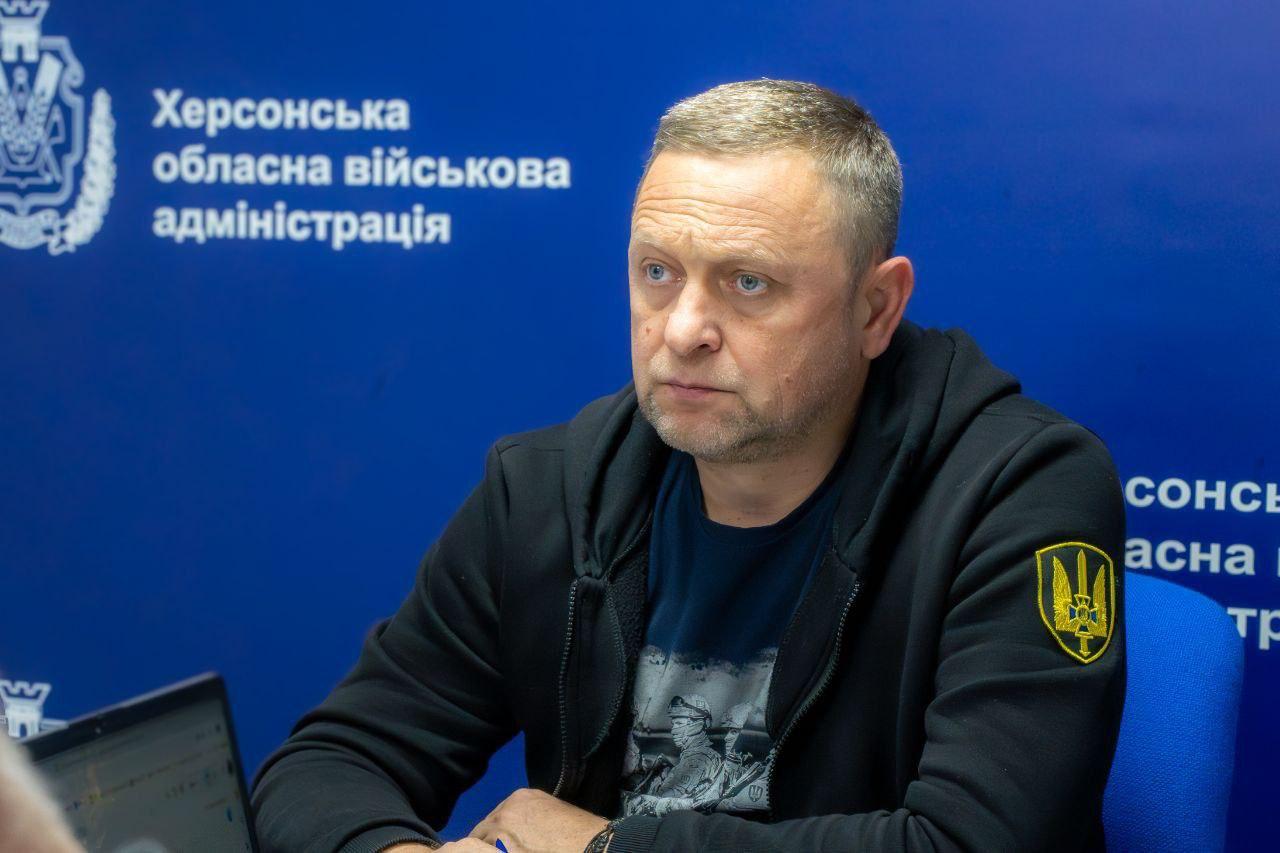 Середній план чоловіка у чорній кофті з капюшоном, який сидить за столом під час наради. На синьому фоні за ним видно напис Херсонська обласна військова адміністрація та герб України на одязі.