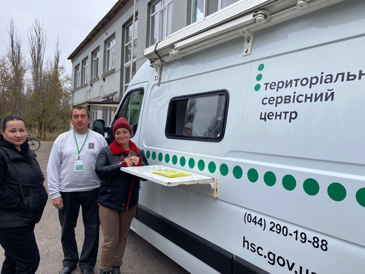 Троє людей (дві жінки та один чоловік) стоять поруч із мобільним Територіальним сервісним центром (ТСЦ) МВС, що виглядає як білий фургон. На фургоні видно напис: територіальний сервісний центр, зелені декоративні елементи, контактний телефон (044) 290-19-88 та веб-адресу: hsc.gov.ua. На задньому плані — сіра будівля та осінні дерева.