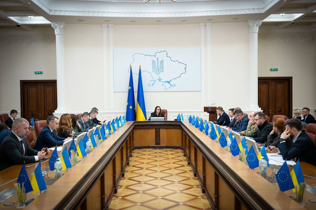 Широкий план довгого столу для засідань в урядовій залі. По центру сидить жінка в чорному костюмі та окулярах. На стіні за нею — велика мапа України з Тризубом. Стіл прикрашений численними маленькими прапорцями України та Європейського Союзу, за ним сидять учасники наради.