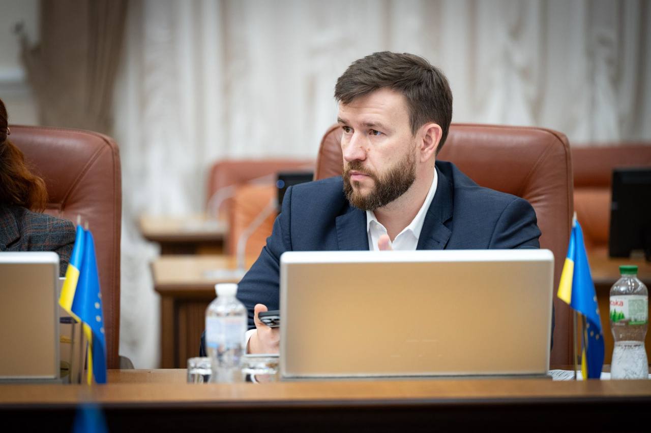 Портретний знімок чоловіка середнього віку з бородою, одягненого у темно-синій піджак та білу сорочку, який сидить за столом на нараді. Перед ним стоїть відкритий ноутбук, пляшка води та мініатюрні прапорці України та Європейського Союзу. Він дивиться вбік із серйозним виразом обличчя.