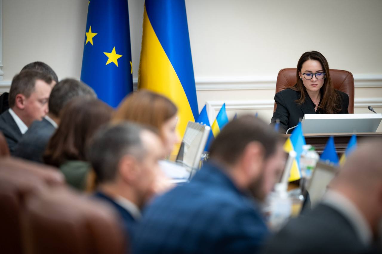 Жінка в окулярах та чорному костюмі сидить за столом під час зустрічі, дивлячись прямо вперед. На задньому плані видно прапори України та Європейського Союзу. Навколо неї — присутні наради, розмиті на передньому плані.