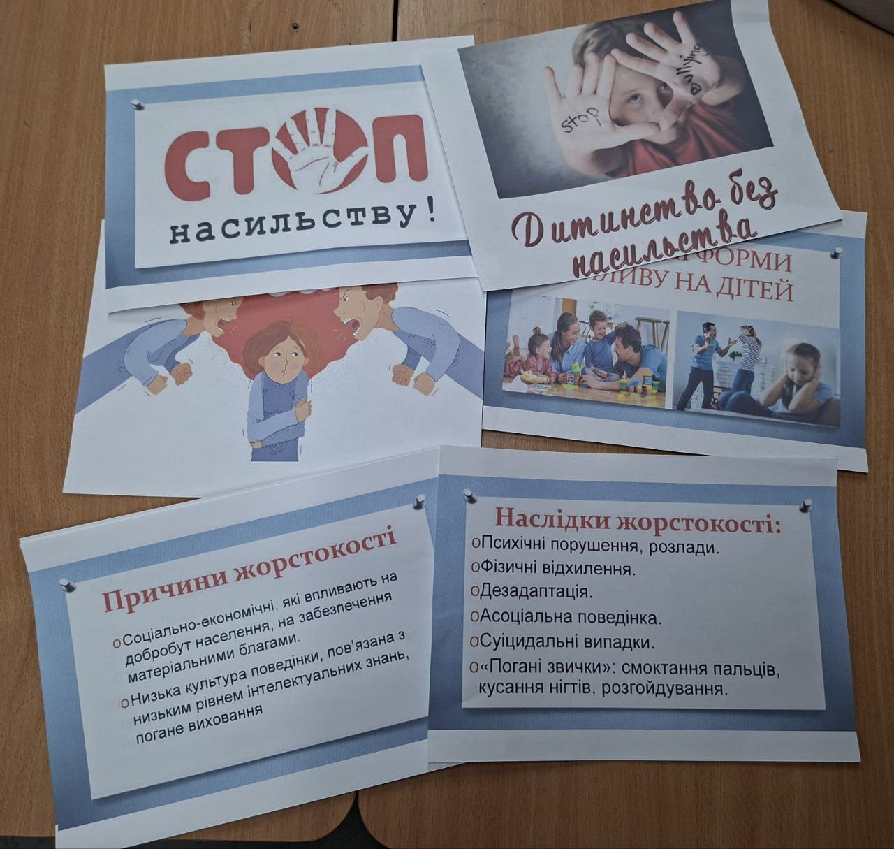 Крупний план розкладених інформаційних плакатів на тему боротьби з насильством та жорстокістю. Видно заголовки: СТОП НАСИЛЬСТВУ!, Дитинство без насильства, Причини жорстокості та Наслідки жорстокості. Текст описує соціально-економічні та культурні причини жорстокості, а також її психологічні, фізичні та соціальні наслідки.
