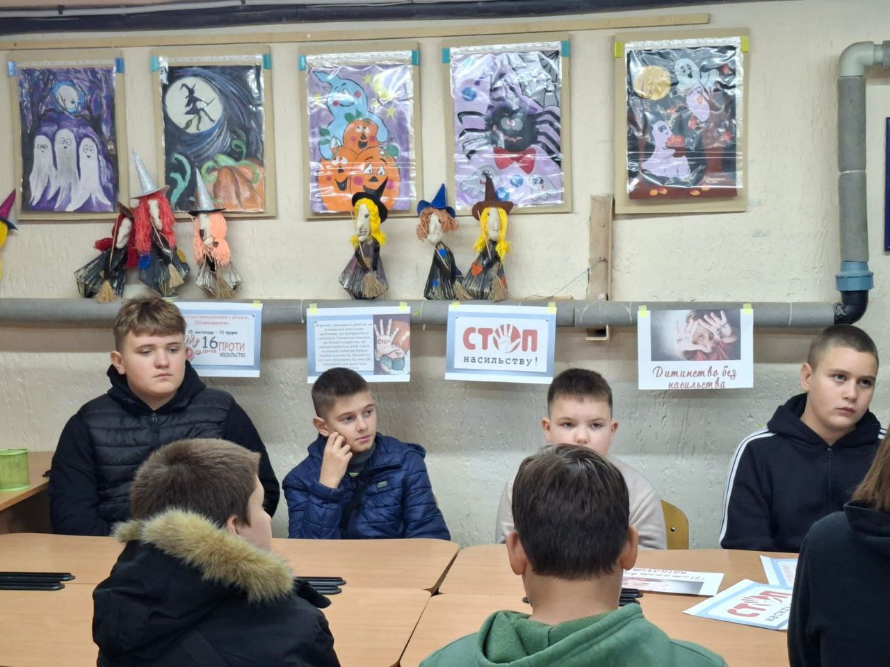 Група підлітків (хлопців) сидить за столом під час виховного заходу чи уроку. Над ними на стіні висять тематичні плакати та малюнки, імовірно, присвячені Гелловіну, а також інформаційні таблички із закликами проти насильства, зокрема СТОП НАСИЛЬСТВУ!