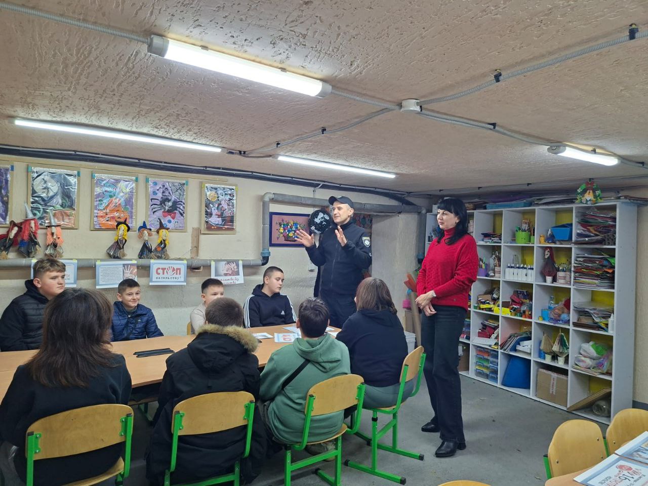 Поліцейський у формі проводить лекцію або виховну бесіду для групи підлітків, які сидять за столом. Поруч стоїть жінка у червоному светрі. Захід, імовірно, відбувається у шкільному приміщенні або укритті, на що вказують низька стеля та стелажі зліва. На стіні висять дитячі малюнки та плакати, зокрема СТОП НАСИЛЬСТВУ!