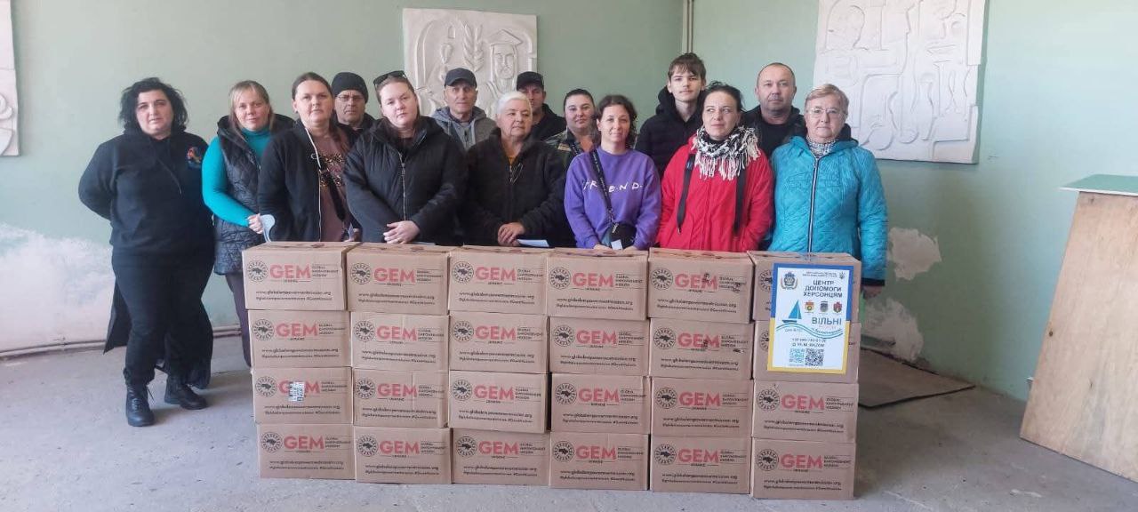 Групова фотографія, на якій близько 14 дорослих людей стоять, позуючи за великою кількістю складених картонних коробок з гуманітарною допомогою від організації GEM (Global Empowerment Mission). Збоку видно інформаційний плакат Центр допомоги Херсонцям організації Вільні Разом. Люди одягнені в теплий повсякденний одяг.