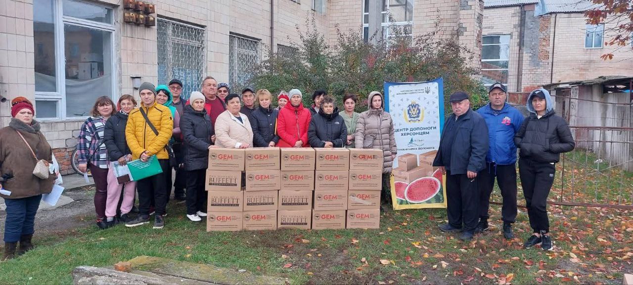 Групова фотографія людей, які стоять на вулиці перед стопкою коробок з гуманітарною допомогою від GEM та Howard G. Buffett Foundation. Поруч з коробками стоїть банер Центр допомоги Херсонцям з логотипом Херсонської ОВА та зображенням кавуна. Група з близько 20 осіб, одягнених у теплий одяг, позує біля допомоги на тлі будівлі та осінньої рослинності.