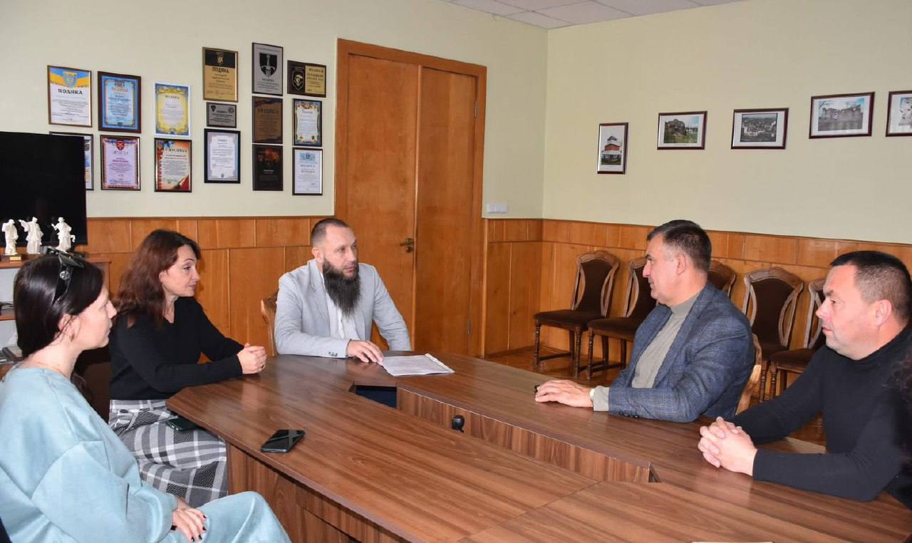 П'ятеро людей (дві жінки та троє чоловіків) сидять за великим дерев'яним столом під час ділової зустрічі чи наради. Чоловік у світло-сірому піджаку та з бородою, що сидить по центру, тримає документи. На стіні позаду них висить багато рамок із дипломами, нагородами та невеликими фотографіями.