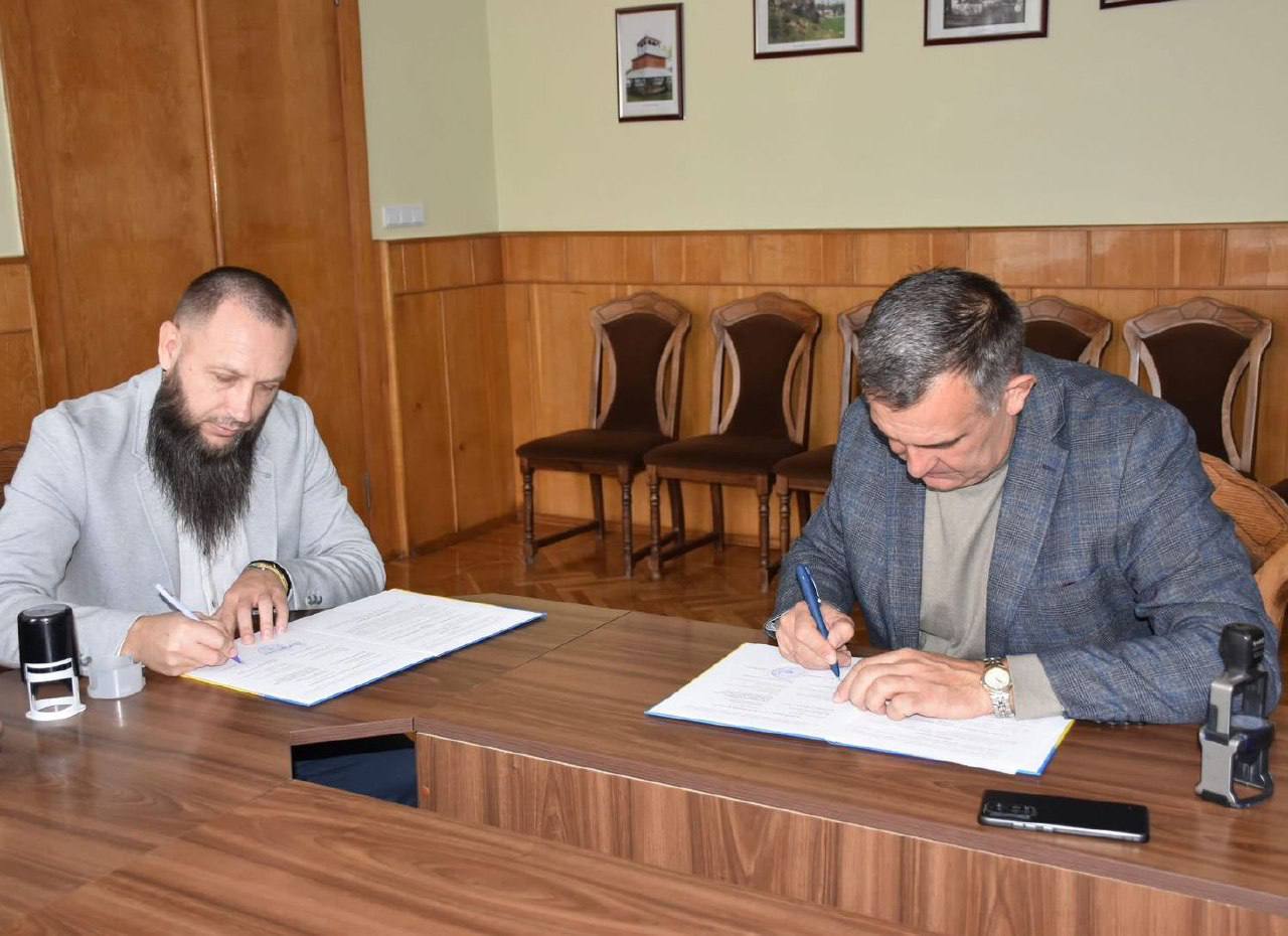 Два чоловіки, які сидять за дерев'яним столом, підписують документи. Чоловік зліва, з довгою бородою і в світло-сірому піджаку, і чоловік справа, у синьому картатому піджаку, одночасно ставлять свої підписи на аркушах паперу. Це ілюструє момент укладення угоди чи підписання договору.