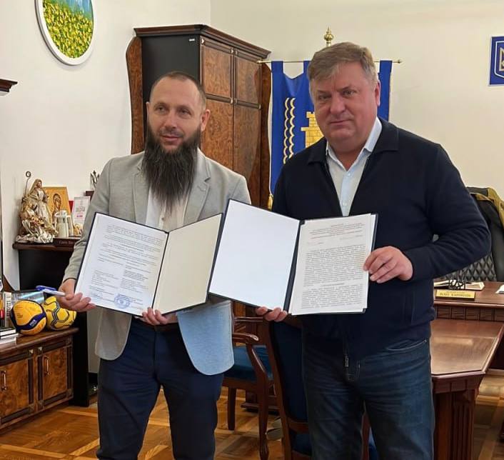 Двоє чоловіків у діловому одязі стоять у приміщенні, схожому на офіс, і тримають відкриті документи. Чоловік ліворуч має бороду і одягнений у світлий піджак, а чоловік праворуч у темно-синьому светрі.