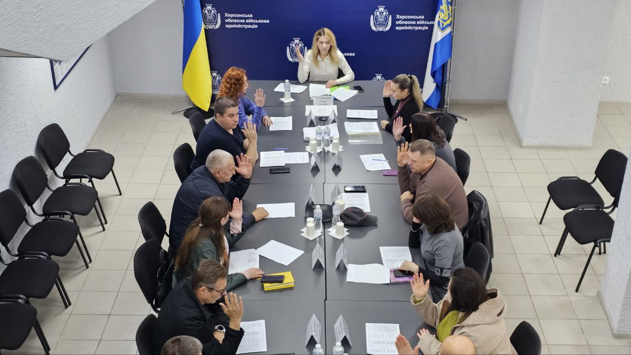 Група людей сидить за довгим столом у конференц-залі, ймовірно, під час засідання в Херсонській обласній військовій адміністрації. Більшість учасників підняли руки вгору для голосування. На тлі видно прапори України та Херсонської ОВА, а також її логотипи.