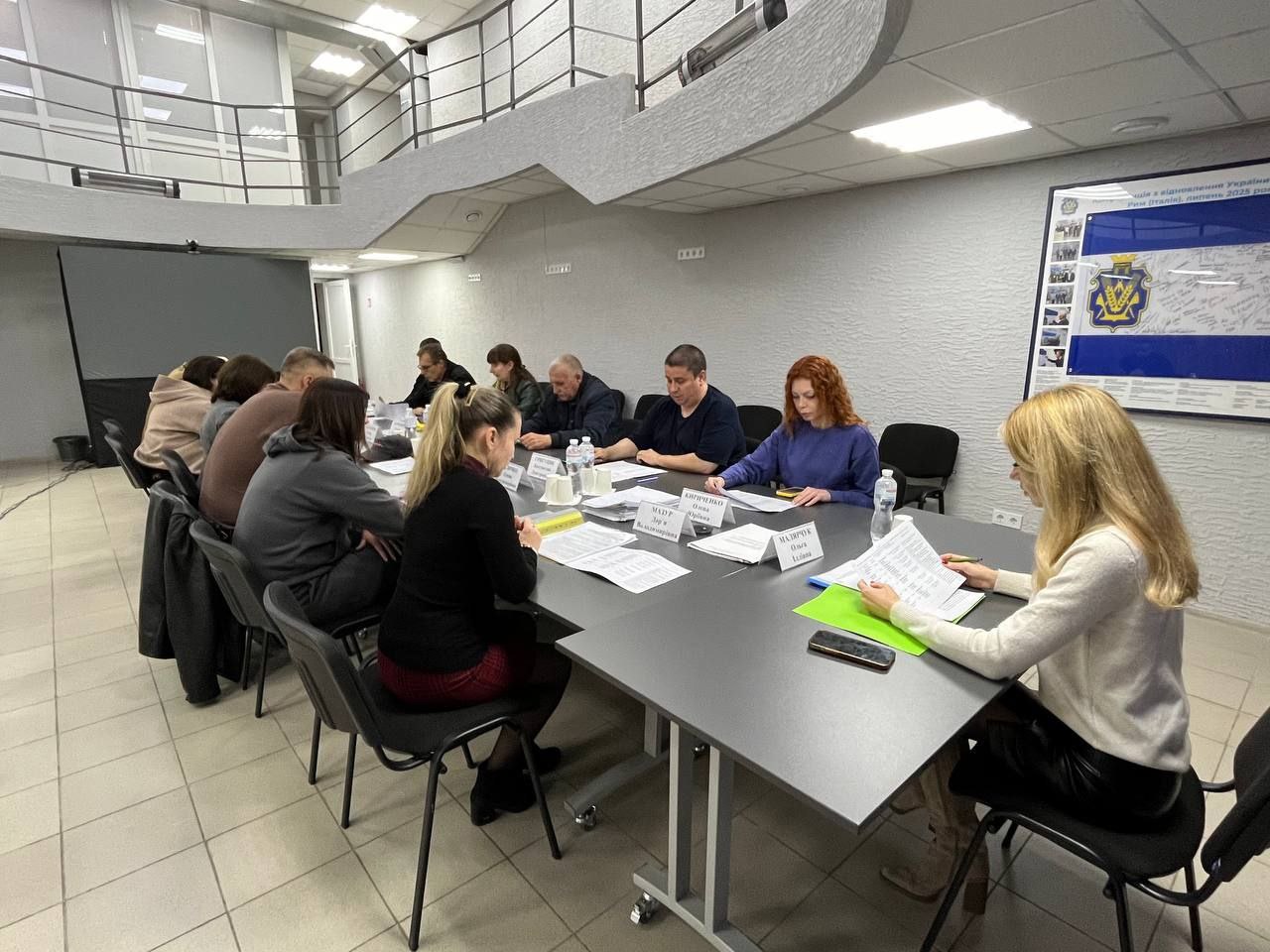 Група людей сидить за довгим столом на діловій зустрічі чи нараді в приміщенні з сірими стінами. Учасники зустрічі переглядають документи, що лежать на столі, деякі з яких мають іменні таблички. На стіні праворуч висить великий плакат з логотипом Херсонської обласної військової адміністрації.