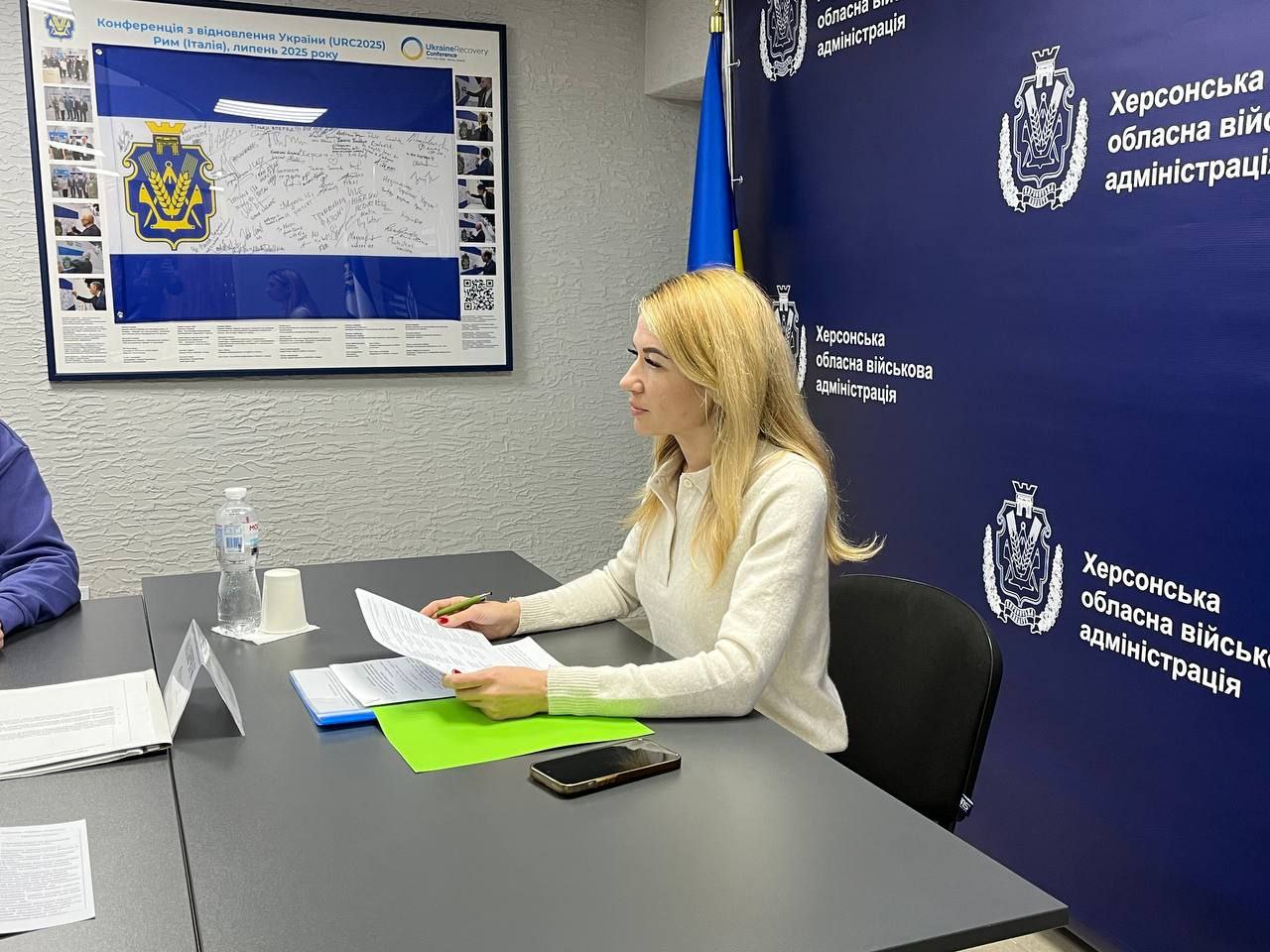 Жінка зі світлим волоссям сидить за столом і тримає документи під час наради. Вона одягнена у світлий светр. На задньому плані розташовані сині банери з написом Херсонська обласна військова адміністрація та її логотипом.