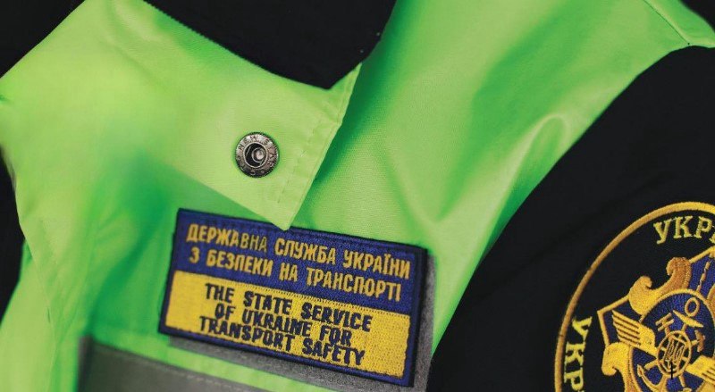 Крупний план нашивки на яскраво-салатовій (жовто-зеленій) світловідбивній формі. На нашивці синього та жовтого кольорів нанесено текст: ДЕРЖАВНА СЛУЖБА УКРАЇНИ З БЕЗПЕКИ НА ТРАНСПОРТІ та нижче англійською: THE STATE SERVICE OF UKRAINE FOR TRANSPORT SAFETY. Поруч видно частину круглої емблеми.