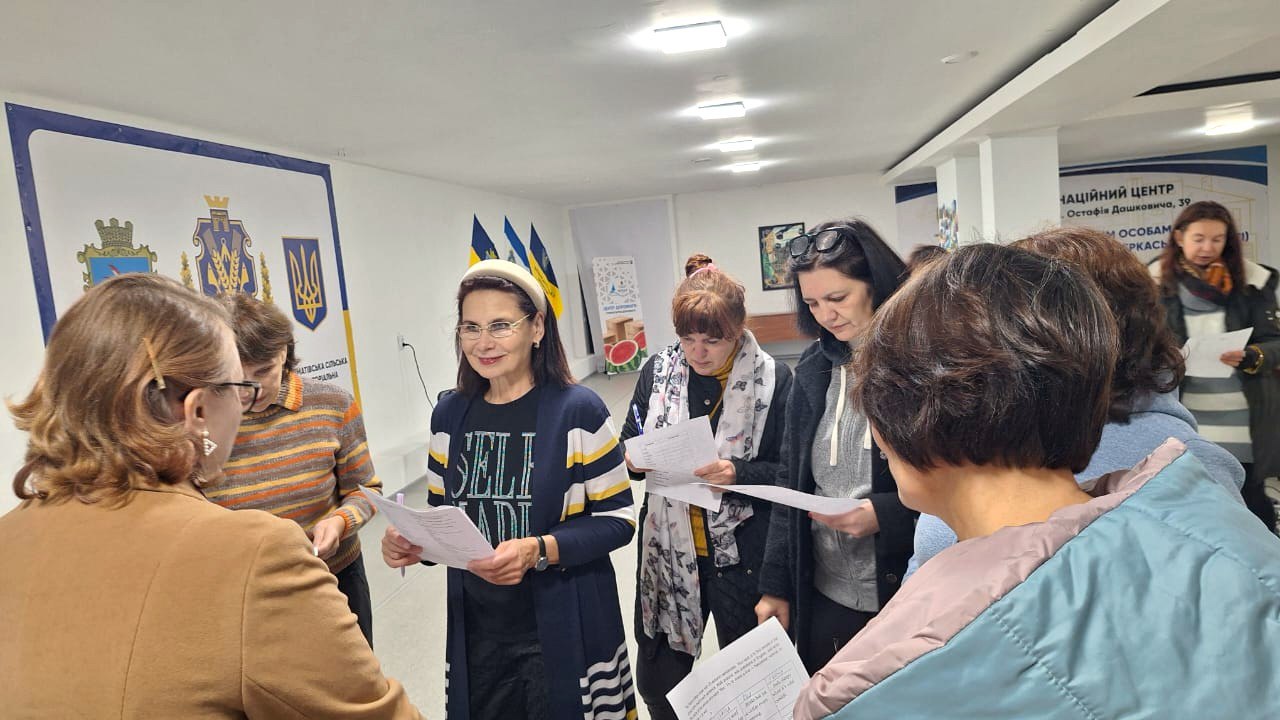 Група жінок стоїть у приміщенні та спілкується, тримаючи в руках аркуші паперу, ймовірно, під час реєстрації або неформальної частини заходу. На задньому плані видно банери з символікою Херсонської обласної військової адміністрації (герби та прапори України і Херсонської області), а також інформацію про Національний центр.