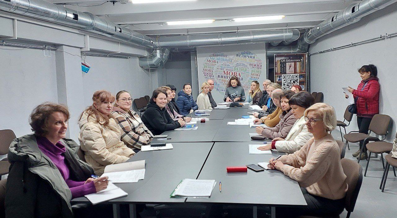 Група з приблизно 20 дорослих людей, переважно жінок, сидить за довгим прямокутним столом у приміщенні з сірими стінами та відкритими повітроводами на стелі. Вони беруть участь у нараді чи освітньому заході. На задньому плані видно банер зі словами, пов'язаними з українською мовою та культурою.