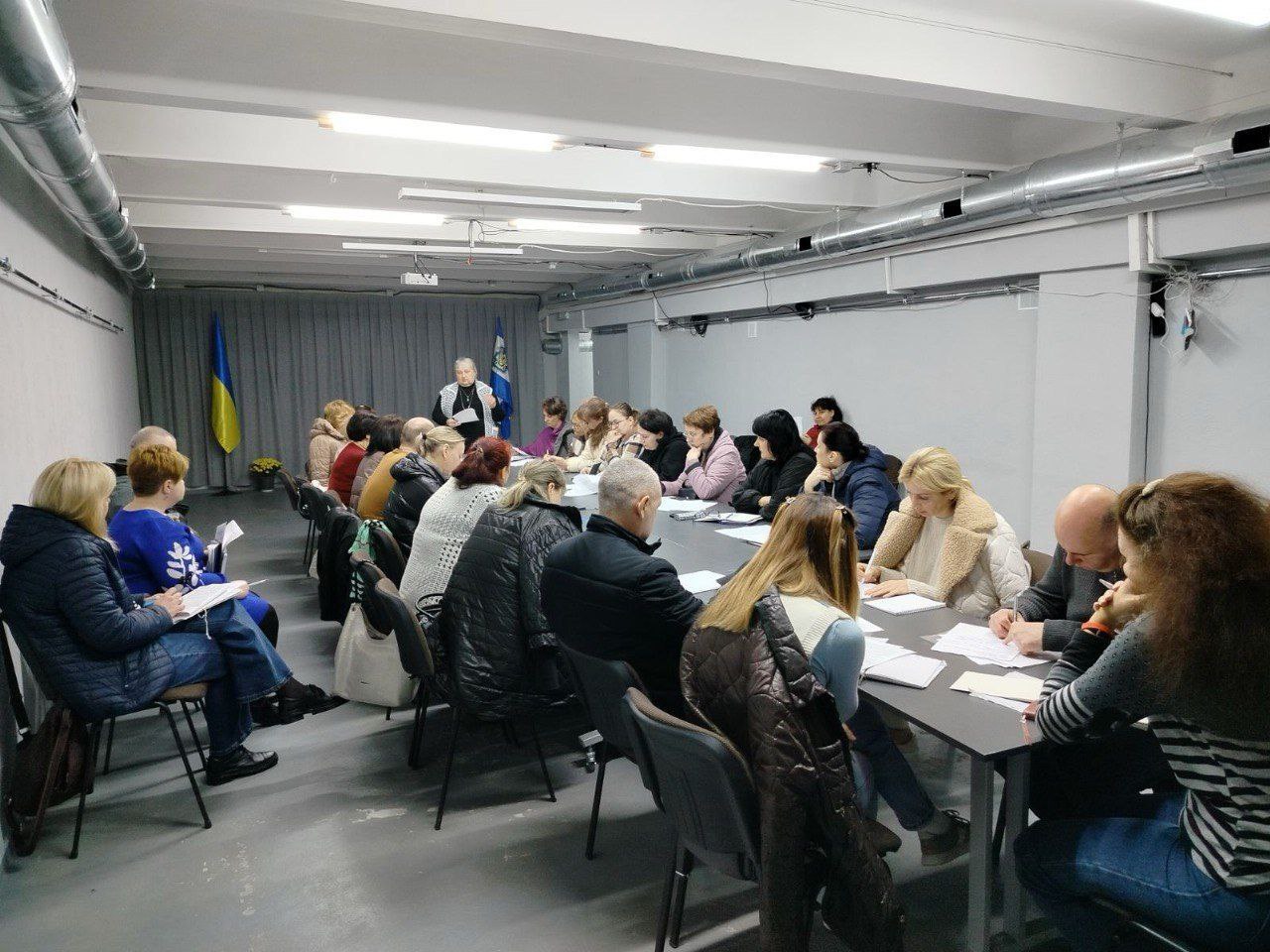 Широкий план зібрання дорослих людей за довгим столом у мінімалістичному приміщенні. Учасники слухають жінку, що стоїть на чолі столу. На задньому плані видно український прапор та інший, ймовірно, місцевий прапор.