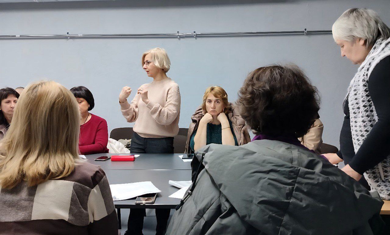 Крупний план групи дорослих людей, які беруть участь у нараді чи обговоренні. У центрі блондинка у світлому светрі стоїть, емоційно жестикулюючи, звертаючись до присутніх за столом.