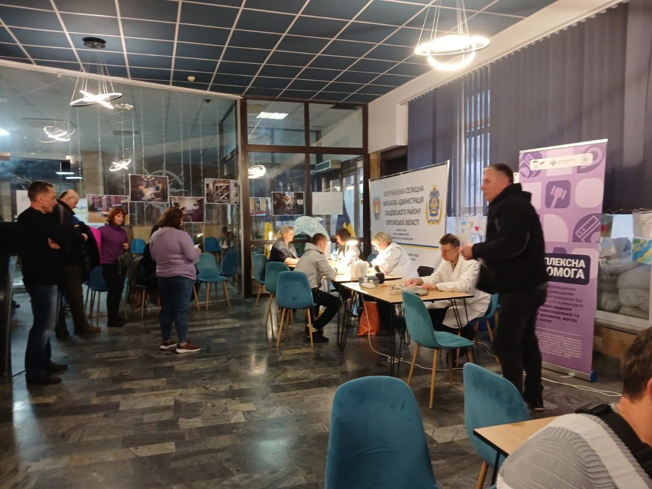 Група людей у приміщенні, схожому на вестибюль, де організовано пункт допомоги. За столами сидять люди у білих халатах, які надають послуги чи консультації іншим відвідувачам. Видно банер з написом: Комплексна допомога та символікою Каховської районної військової адміністрації.