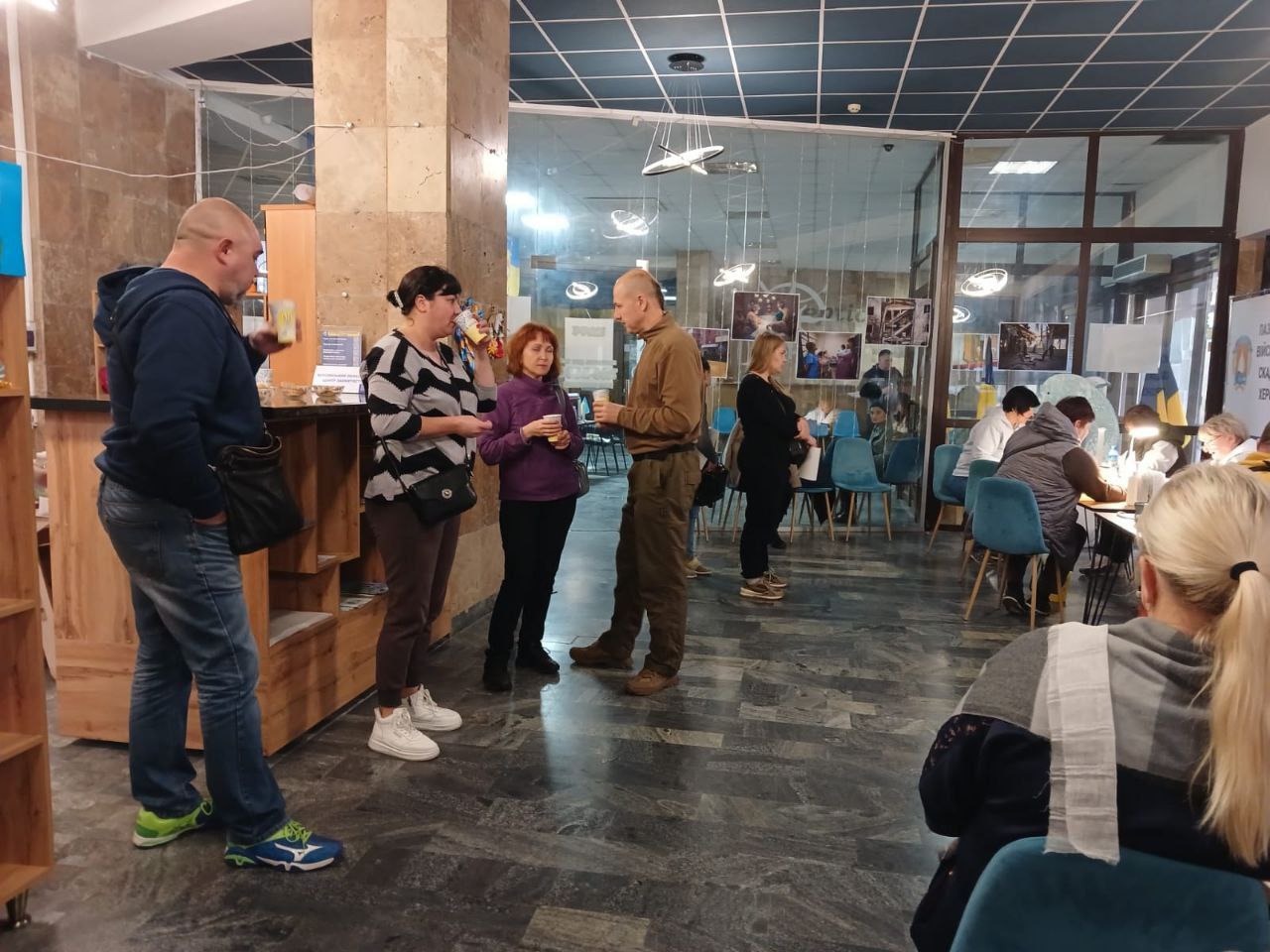 Кілька груп людей стоять і спілкуються у великому сучасному приміщенні з високою стелею. На задньому плані видно столи, за якими відбувається надання послуг в рамках гуманітарного та соціального заходу, що відповідає попередньому зображенню.