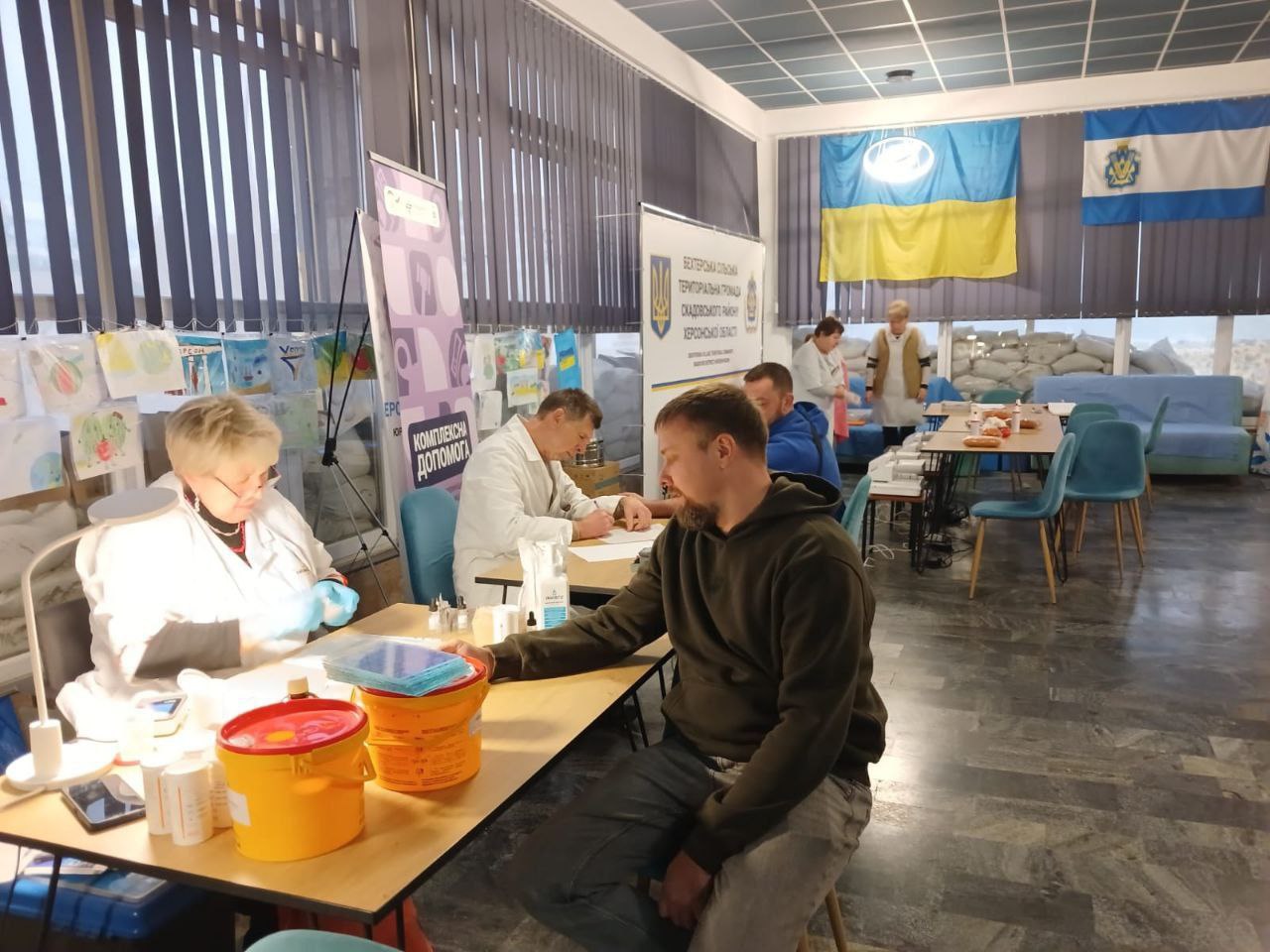 Сцена з медичного пункту, розташованого у приміщенні. Жінка у білому халаті працює за столом, перед нею сидить чоловік. Позаду видно український прапор та прапор Каховського району, а також мішки з піском, що вказує на прифронтову або небезпечну зону. На банері читається напис: Територіальна громада Каховського району Херсонської області.