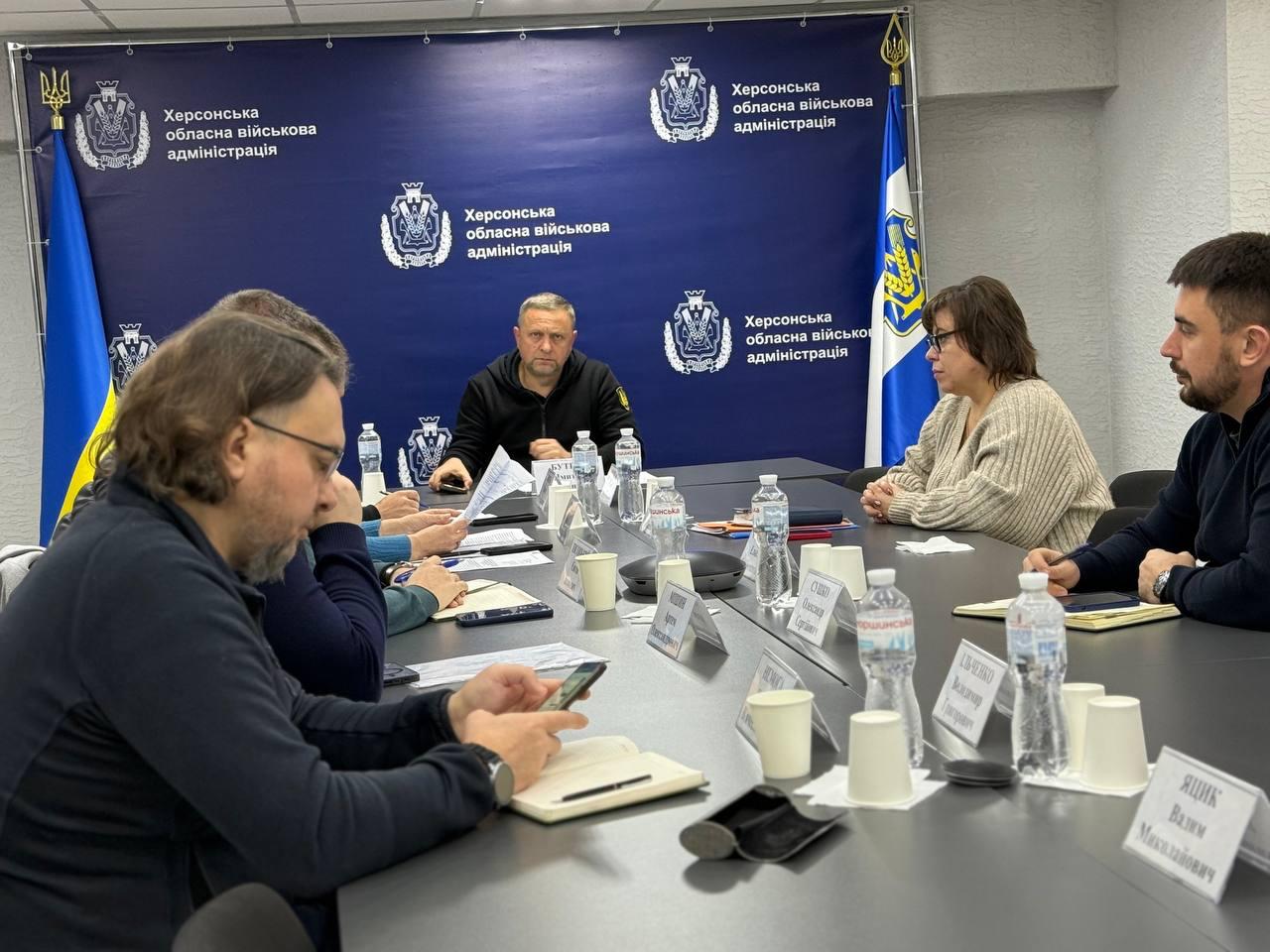 На зображенні — засідання за великим столом у приміщенні Херсонської обласної військової адміністрації (про що свідчить напис на банері позаду). Учасники, серед яких чотири людини чітко видно (троє чоловіків та одна жінка), сидять навколо столу, заставленого пляшками води, склянками та табличками з іменами. Центральна особа (чоловік у темній військовій куртці) дивиться вперед, тримаючи папери. Один з учасників ліворуч дивиться у свій телефон.