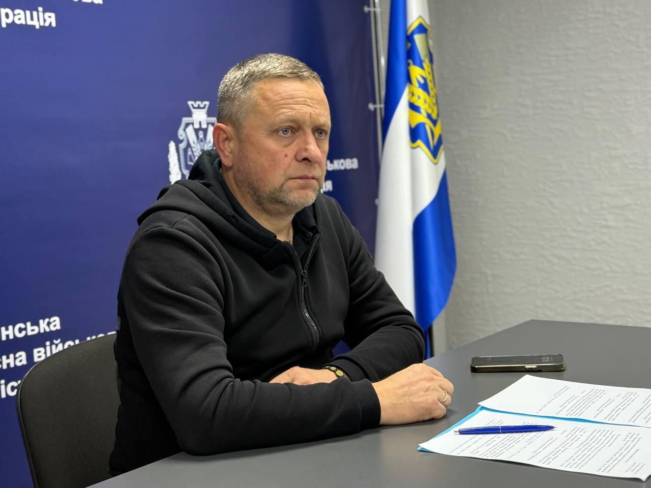 На фотографії зображено чоловіка середнього віку з коротким сивіючим волоссям, одягненого у чорну кофту на блискавці (худі або спортивна куртка), який сидить за столом. Його руки складені перед ним на столі, поруч лежать документи. Фон складається з темно-синього банера та гербом, а також українським прапором та прапором Херсонської області праворуч. Чоловік має серйозний вираз обличчя і дивиться трохи ліворуч від камери.