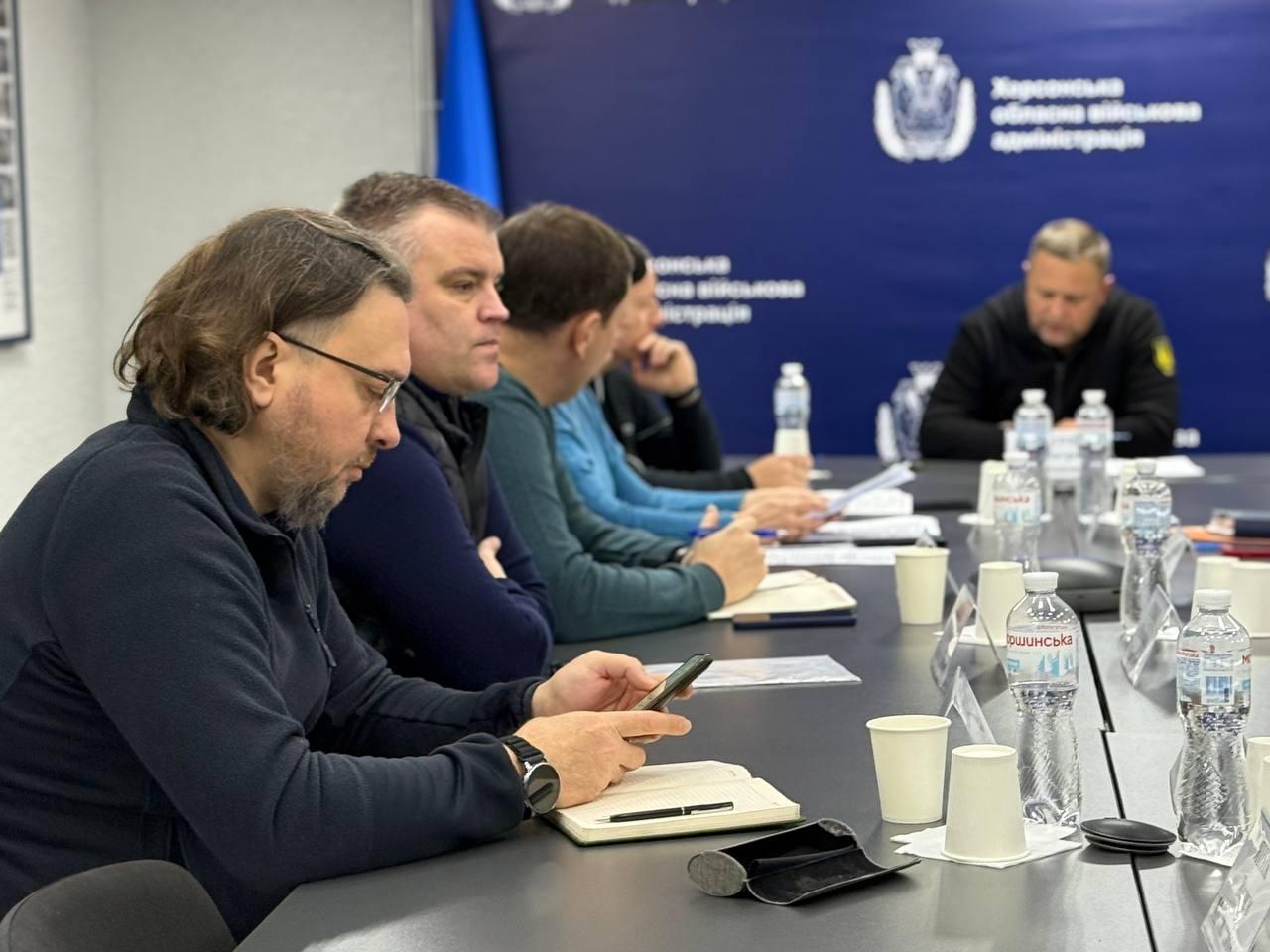 На фотографії зображено нараду за довгим столом. Зліва на передньому плані — чоловік у темному одязі та окулярах, який дивиться у свій смартфон, тримаючи його над відкритим блокнотом. Праворуч від нього сидять ще кілька чоловіків. На задньому плані, частково в тіні, сидить центральна фігура (можливо, голова адміністрації) у чорній кофті, якого було видно на попередніх знімках. На синьому банері на стіні позаду видно написи: Херсонська обласна військова адміністрація та герб.