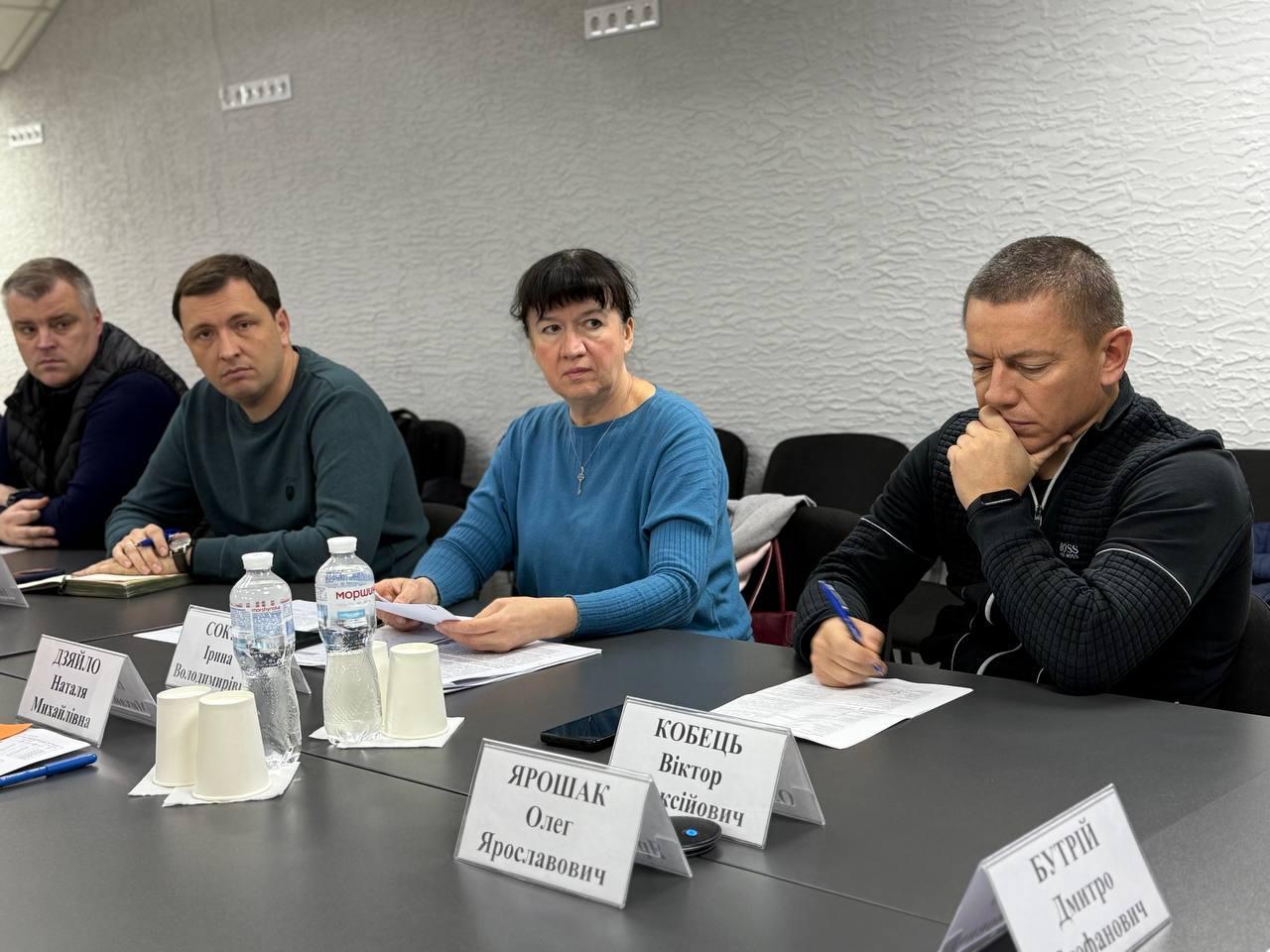 На фотографії — четверо учасників (троє чоловіків та одна жінка) сидять за столом під час наради. Жінка в центрі в блакитному светрі тримає папери. Чоловік праворуч (у чорній спортивній куртці з логотипом BOSS) схилився над паперами, щось записуючи. На столі видно таблички з іменами та прізвищами учасників. Фон — світла рельєфна стіна.