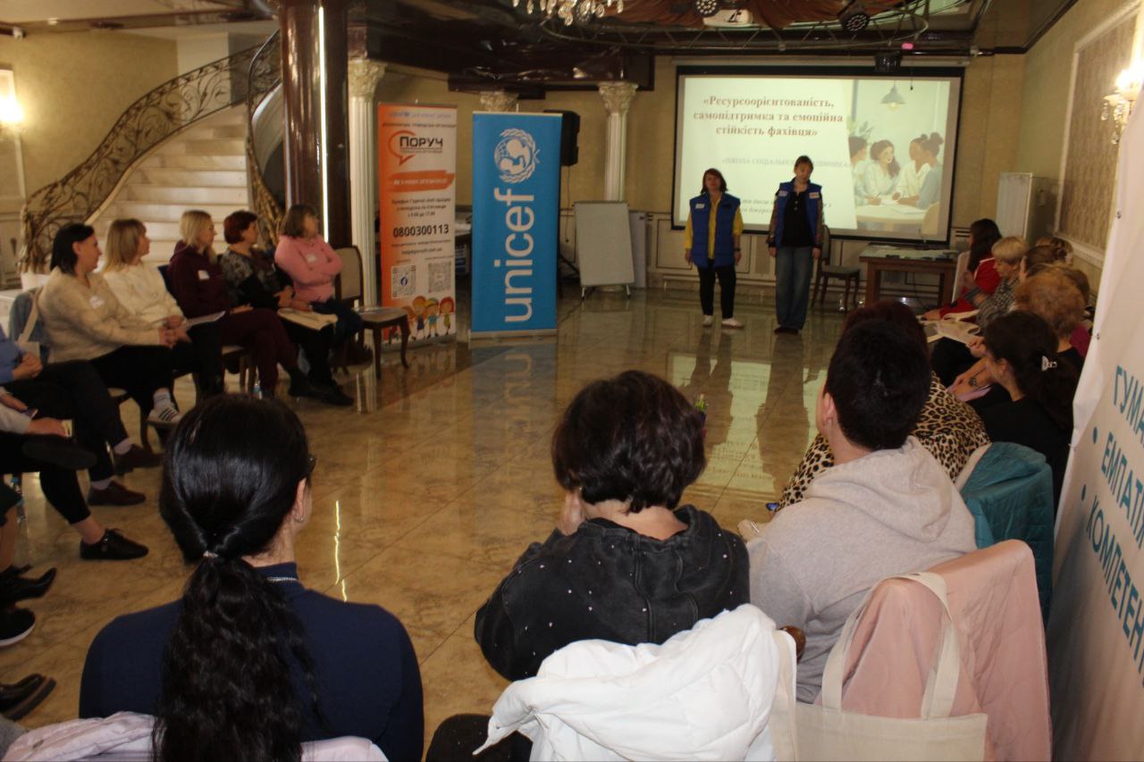 Учасники сидять у приміщенні та слухають двох жінок, які стоять біля екрана з презентацією на тему: Ресурсоорієнтованість, самопідтримка та емоційна стійкість фахівця, поруч видно банери: ЮНІСЕФ та ПОРУЧ.