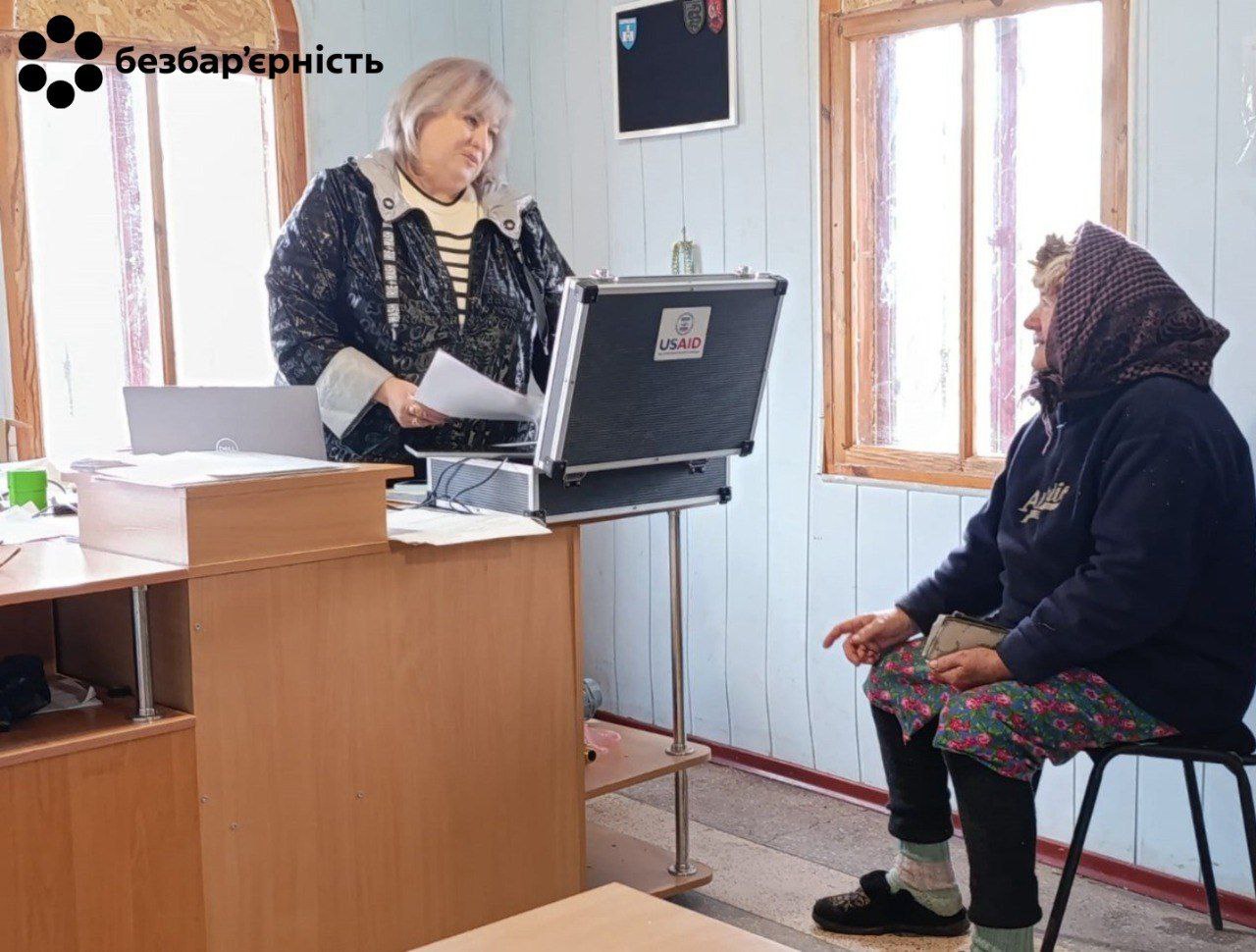 Дві жінки в приміщенні біля дерев'яного столу: одна жінка (співробітниця) стоїть за столом, тримаючи документи, поруч стоїть відкритий кейс із логотипом USAID; інша жінка (місцева жителька) сидить на стільці навпроти. У верхньому лівому куті — логотип Безбар'єрність.