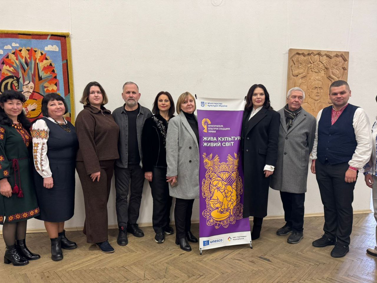 Група з дев'яти осіб, включаючи чоловіків і жінок, стоїть у приміщенні з витворами мистецтва на стінах, позуючи біля фіолетового банера Міністерства культури України з написом: НЕМАТЕРІАЛЬНА КУЛЬТУРНА СПАДЩИНА УКРАЇНИ. ЖИВА КУЛЬТУРА ЖИВИЙ СВІТ.