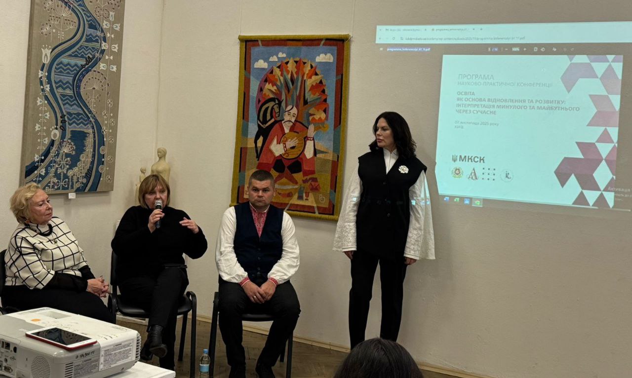 Панельна дискусія або виступ чотирьох осіб у галереї: дві жінки сидять ліворуч, одна з мікрофоном, чоловік у вишиванці сидить по центру, а інша жінка у чорно-білому костюмі стоїть праворуч. На задньому плані видно декоративні килими та проекційний екран зі слайдом: ПРОГРАМА НАУКОВО-ПРАКТИЧНОЇ КОНФЕРЕНЦІЇ.