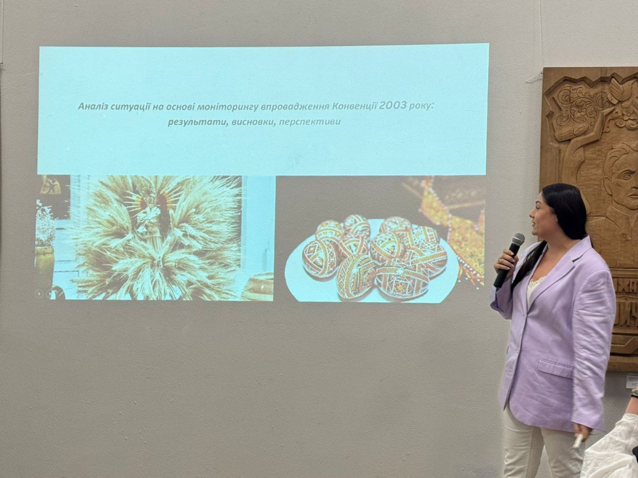 Жінка у світло-бузковому піджаку виступає з мікрофоном, стоячи біля проекційного екрана, на якому відображено слайд з текстом: Аналіз ситуації на основі моніторингу впровадження Конвенції 2003 року: результати, висновки, перспективи та зображеннями виробів народного мистецтва.
