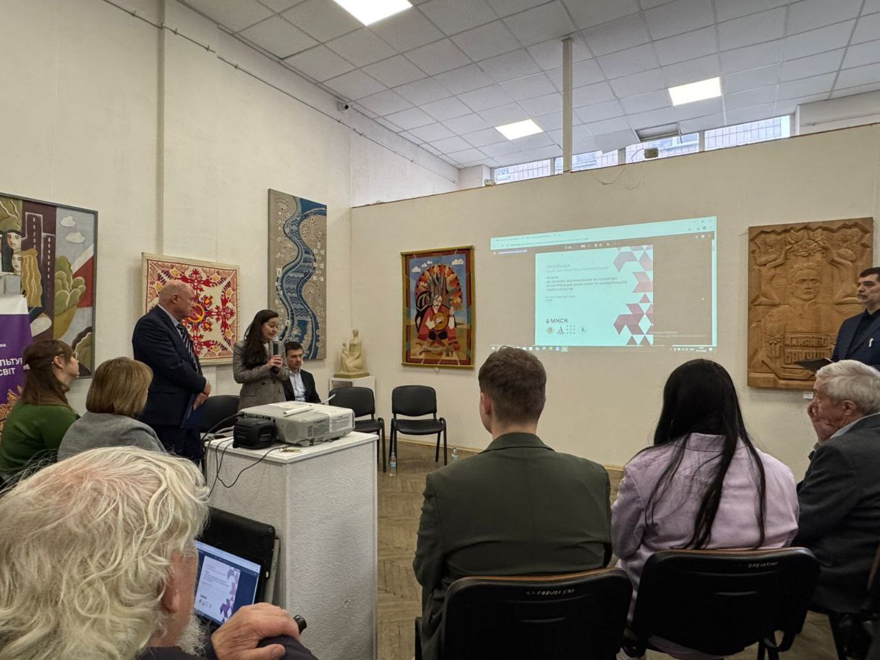 У приміщенні з картинами та виробами мистецтва на стінах відбувається презентація: жінка виступає з мікрофоном, поруч стоїть чоловік, а аудиторія сидить обличчям до них. На екрані видно слайд презентації.