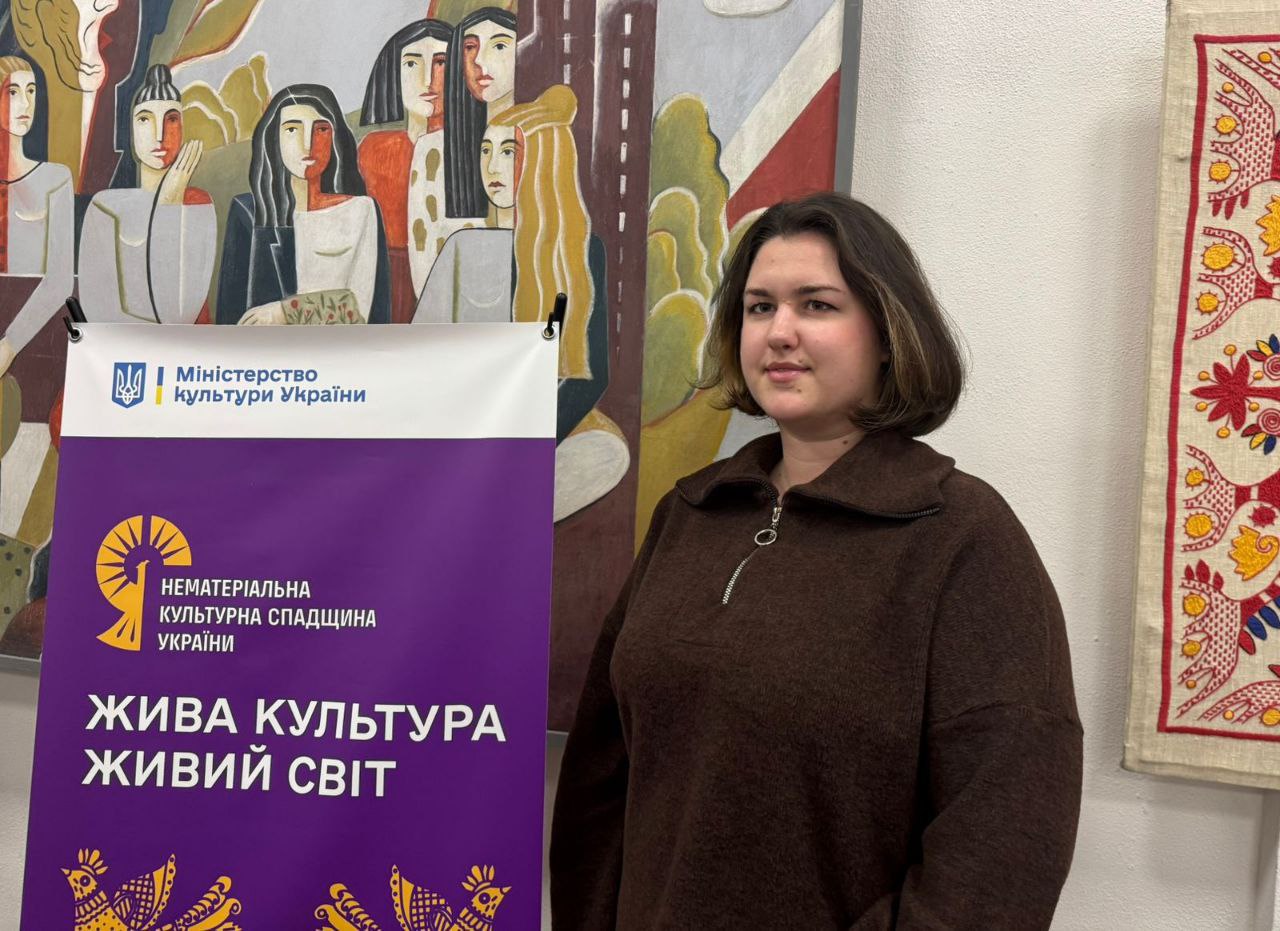 Молода жінка у коричневому светрі стоїть біля банера з логотипом Міністерства культури України та написом: НЕМАТЕРІАЛЬНА КУЛЬТУРНА СПАДЩИНА УКРАЇНИ. ЖИВА КУЛЬТУРА ЖИВИЙ СВІТ. На задньому плані видно картину з фігурами людей та елементи вишивки.