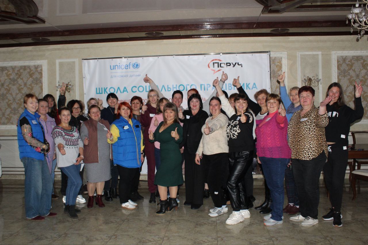 Група з приблизно двадцяти жінок стоїть у приміщенні перед банером: ЮНІСЕФ та ПОРУЧ з написом: ШКОЛА СОЦІАЛЬНОГО ПРАЦІВНИКА та жестикулює піднятими великими пальцями вгору, посміхаючись.
