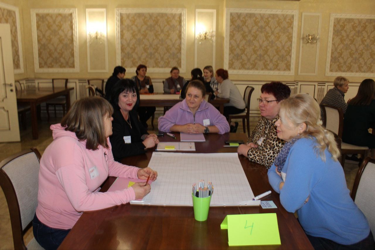 Група з п'яти жінок сидить за великим столом у вишукано оформленій кімнаті з високими стінами, працюючи над великим аркушем паперу. Жінки обговорюють щось у рамках, схожому на семінар або тренінг.