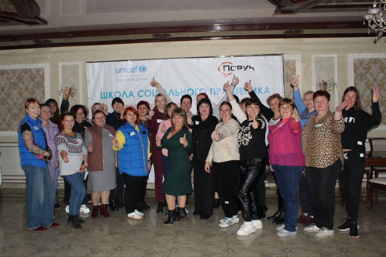 Велика група, що складається переважно з жінок, стоїть і посміхається, показуючи великі пальці вгору (клас або добре) перед банером із логотипами UNICEF та ПОРУЧ. На банері також написано ШКОЛА СОЦІАЛЬНОГО ПРАЦІВНИКА. Люди одягнені у різний повсякденний одяг, ймовірно, після завершення тренінгу чи заходу.