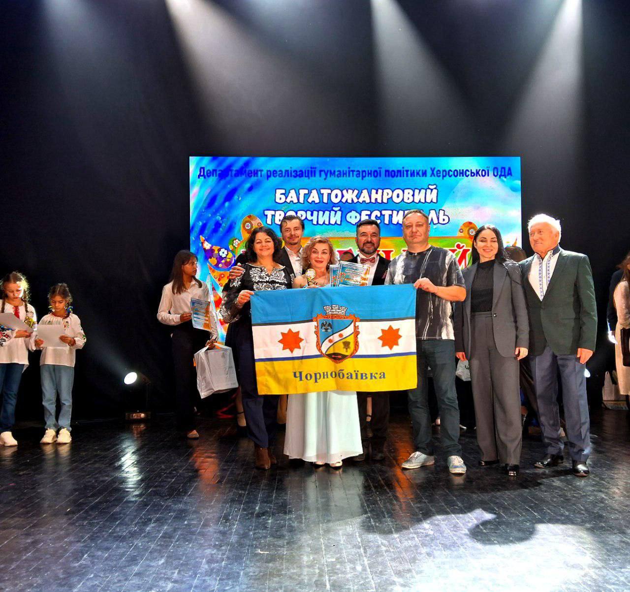 Група з восьми людей стоїть на сцені та тримає прапор із гербом і написом: Чорнобаївка. На задньому плані — екран з написом: Департамент реалізації гуманітарної політики Херсонської ОДА БАГАТОЖАНРОВИЙ ТВОРЧИЙ ФЕСТИВАЛЬ.
