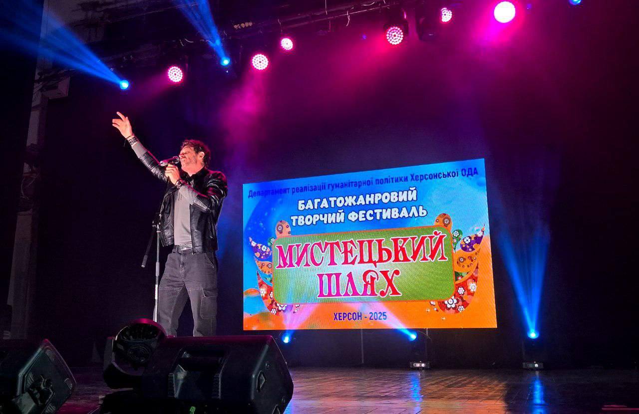 Чоловік співає на сцені з піднятою рукою перед великим екраном з написом: Багатожанровий творчий фестиваль МИСТЕЦЬКИЙ ШЛЯХ ХЕРСОН - 2025 (Департамент реалізації гуманітарної політики Херсонської ОДА).