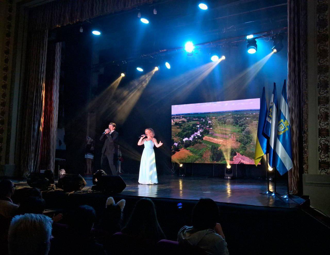 Чоловік у костюмі та жінка у довгій блакитній сукні співають на сцені, освітленій синім світлом, перед екраном зі зображенням сільського пейзажу. Праворуч стоять прапори, схожі на прапори України та Херсонської області.