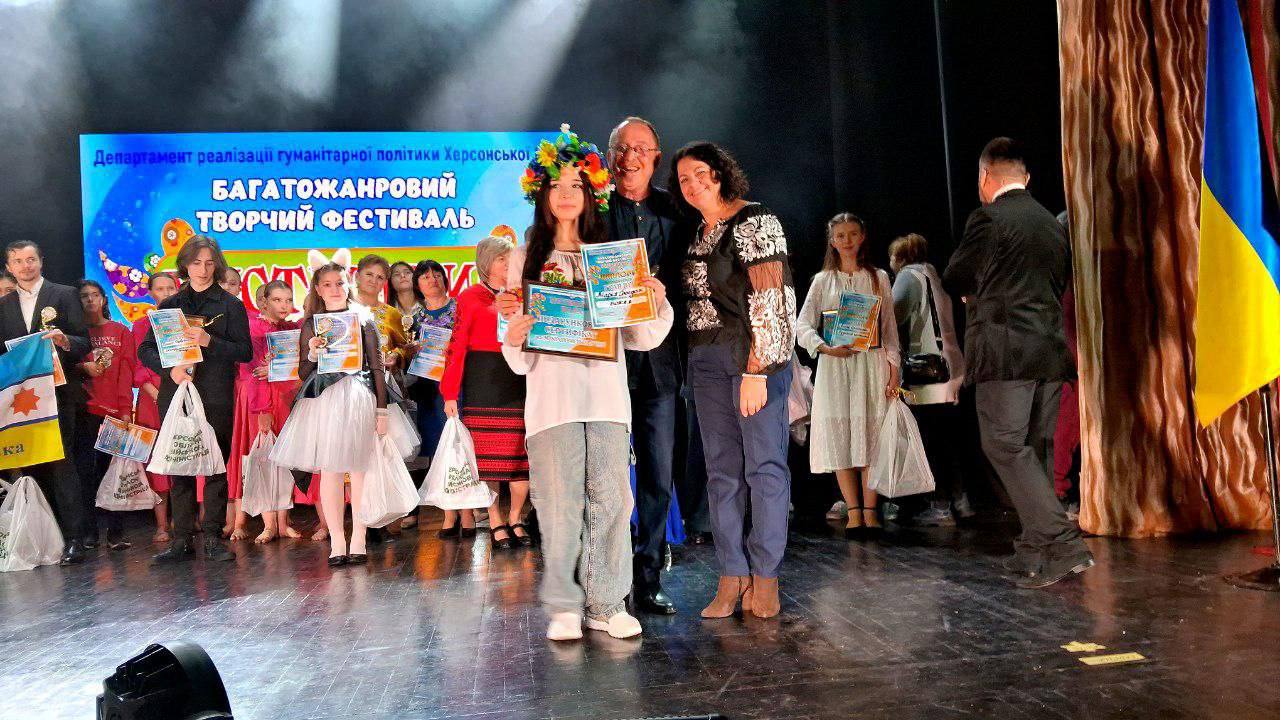 Група людей на сцені, в центрі стоять дівчина у вінку з квітами, яка тримає сертифікати, та двоє дорослих. На задньому плані — великий екран з написом: Департамент реалізації гуманітарної політики Херсонської ОДА БАГАТОЖАНРОВИЙ ТВОРЧИЙ ФЕСТИВАЛЬ. Праворуч видно український прапор.